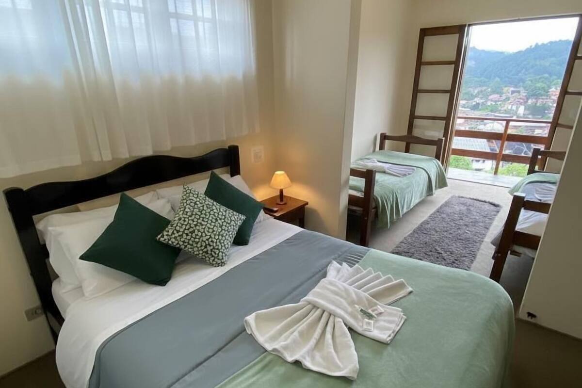 Habitación de hotel con dos camas, una mesita de noche con lámpara y vistas a un paisaje montañoso a través de un ventanal grande.