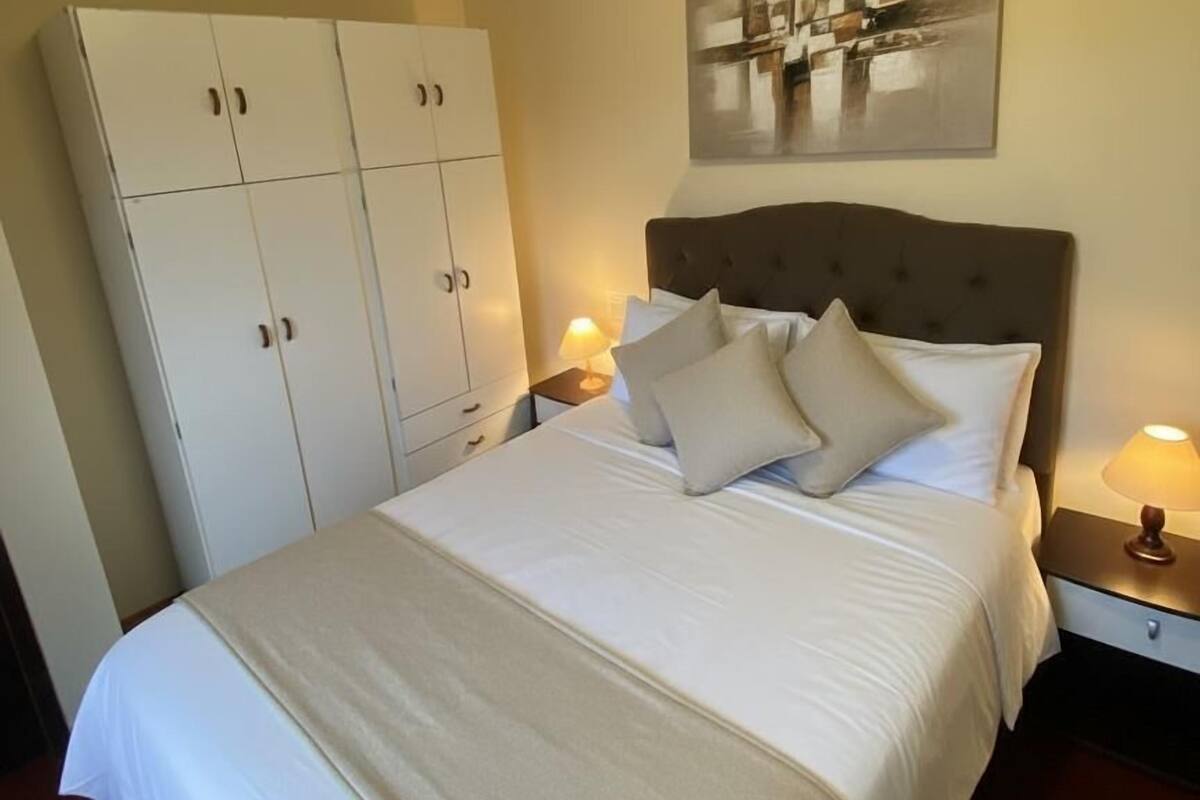 Habitación de hotel con una cama grande, un armario, una mesita de noche con lámpara y un cuadro abstracto colgado en la pared.