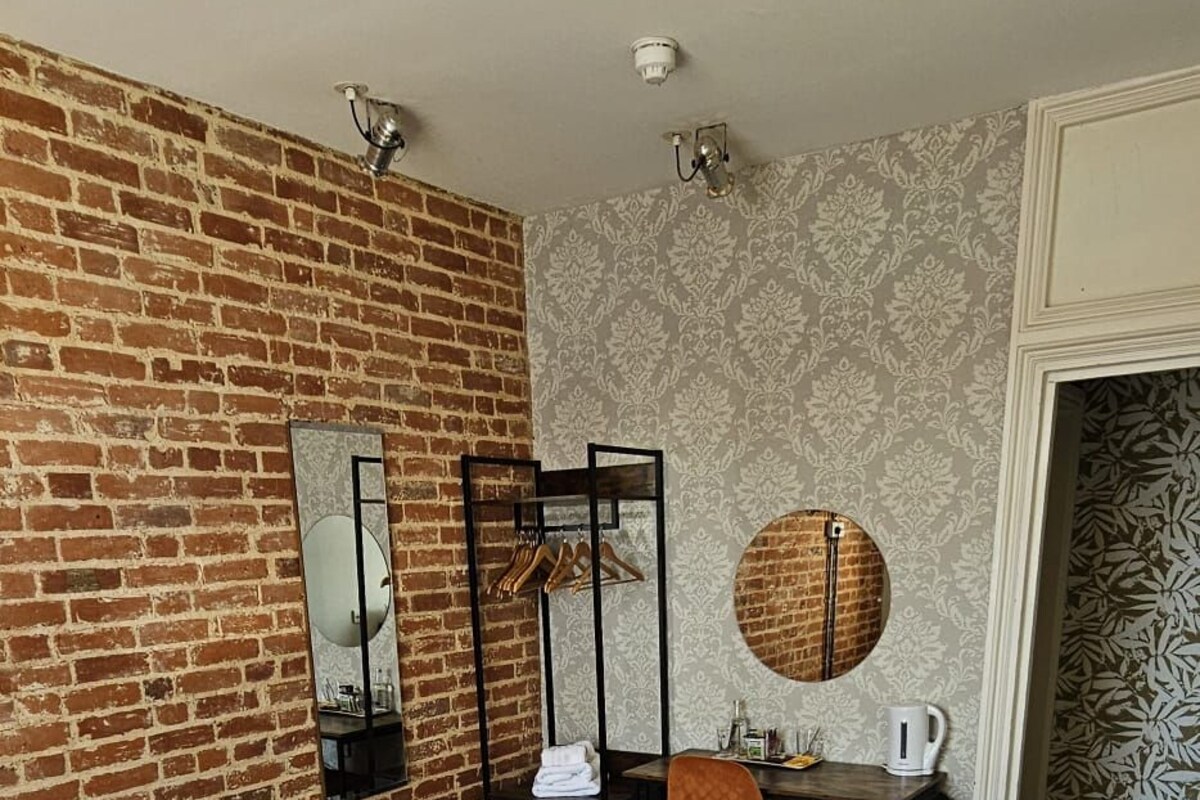 A hotel room with a large bed, a desk with a chair, a hanging coat rack, a round mirror, and a brick wall.