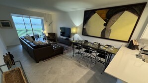 A modern living room with a sofa, a dining table set for a meal, a TV, and a large window overlooking a body of water.