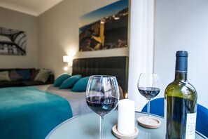 Quarto de hotel com uma cama, uma mesa de vidro com copos de vinho e uma garrafa, e um quadro na parede.