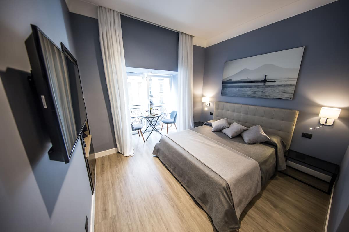 A modern hotel room with a large bed, a flat-screen TV, a balcony with a table and chairs, and a painting of a landscape on the wall.