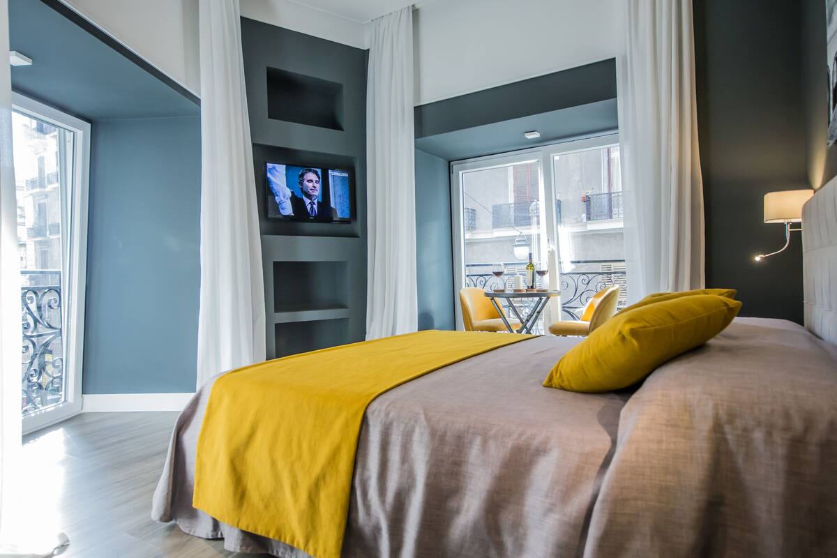A modern hotel room with a large bed, a television, a balcony with a table and chairs, and a city view.