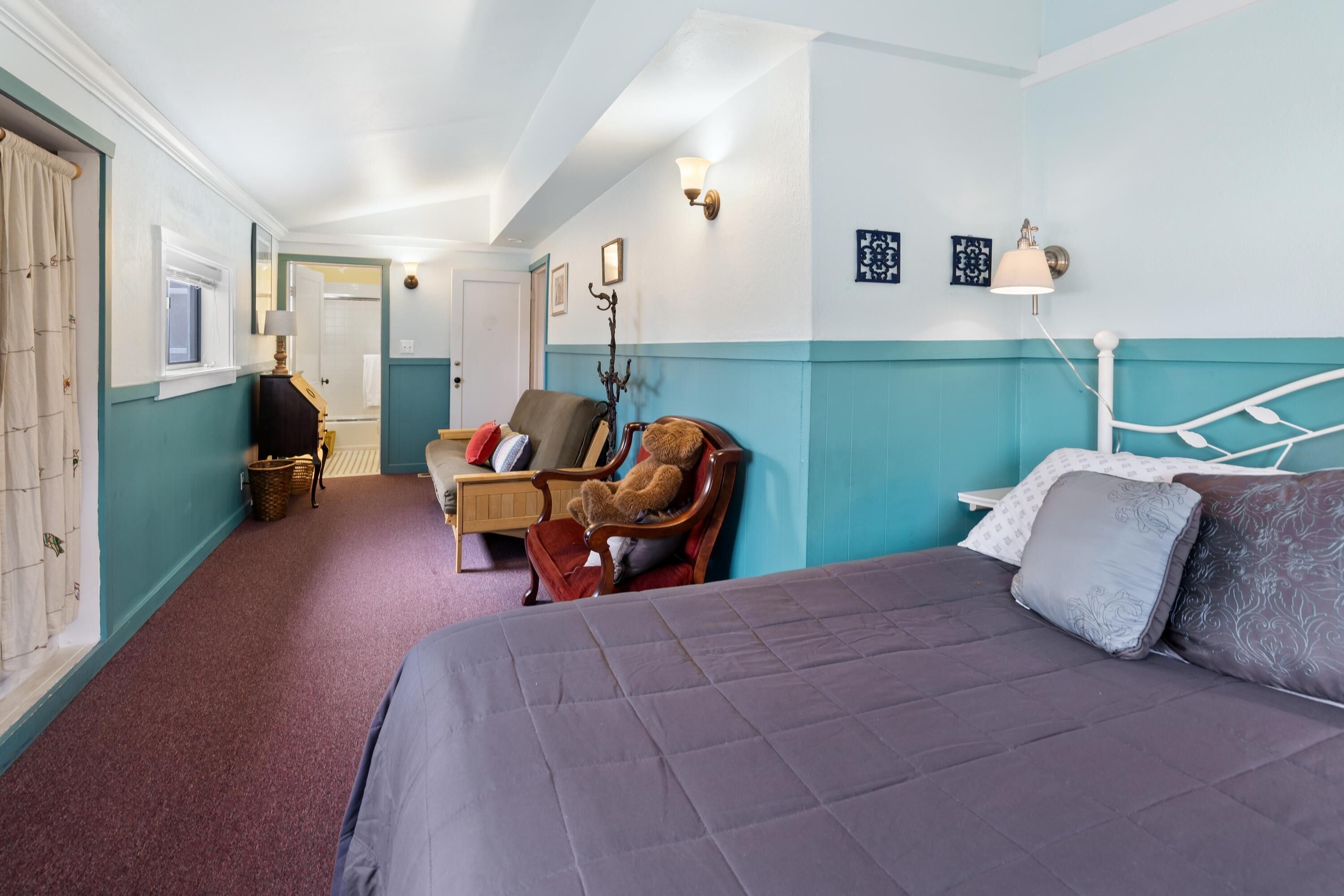 Une chambre d’hôtel avec un lit, une chaise sur laquelle est posé un ours en peluche, une table de chevet et une lampe.