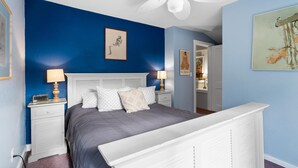 Ein Schlafzimmer mit einem Bett, einem weißen Kopfteil, zwei Nachttischen mit Lampen, einem Deckenventilator und Bildern an den Wänden.
