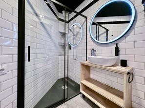 A modern bathroom with white subway tiles, a glass-enclosed shower, a round mirror with a blue light, and a wooden vanity with a white sink. - Charming Madrid Principe Pio (Madrid)