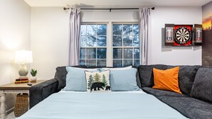A bedroom with a bed, a grey sofa, a window with curtains, a lamp, a small plant, and a dartboard on the wall. - All Utilities! Hot Tub, Lg fenced yard, 10 min to DT, Office, Pets ~ Long Stays! (Asheville)