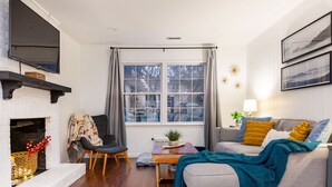 A cozy living room with a fireplace, a sofa, a coffee table, and a TV mounted on the wall. - All Utilities! Hot Tub, Lg fenced yard, 10 min to DT, Office, Pets ~ Long Stays! (Asheville)