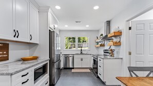 A modern kitchen with white cabinetry, stainless steel appliances, and a wooden dining table. - All Utilities! Hot Tub, Lg fenced yard, 10 min to DT, Office, Pets ~ Long Stays! (Asheville)
