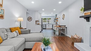 A modern living room with a grey sectional sofa, a wooden coffee table, and a kitchen in the background. - All Utilities! Hot Tub, Lg fenced yard, 10 min to DT, Office, Pets ~ Long Stays! (Asheville)
