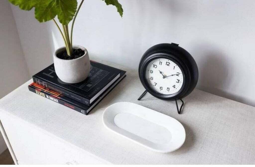 A white table with a black clock, a white plate, and a potted plant on a stack of books.