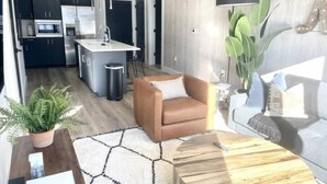 Une cuisine moderne avec des meubles de couleur sombre, un îlot central et un évier. Le salon est aménagé avec un canapé en cuir marron et une table basse ronde en bois.