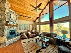 A cozy living room with a stone fireplace, leather furniture, and large windows offering a view of the outdoors.