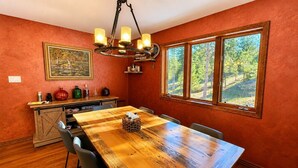 A dining area with a long wooden table, chairs, a chandelier, a window with a view of trees, and a kitchen area with cabinets and a sink. - Sundance & RMNP Escape Backing National Forest (Lyons)