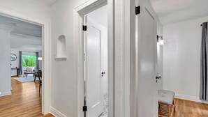 Un passadís amb parets blanques, tarima de fusta i una porta que dona a una habitació amb finestra i vista a vegetació.