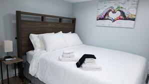 A neatly made bed with white linens, a wooden headboard, and a small bedside table with a lamp and remote control. - Cozy NYC on the Island (West Hempstead)