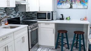 A modern kitchen with white cabinets, a microwave, a stove, and two stools. - Cozy NYC on the Island (West Hempstead)