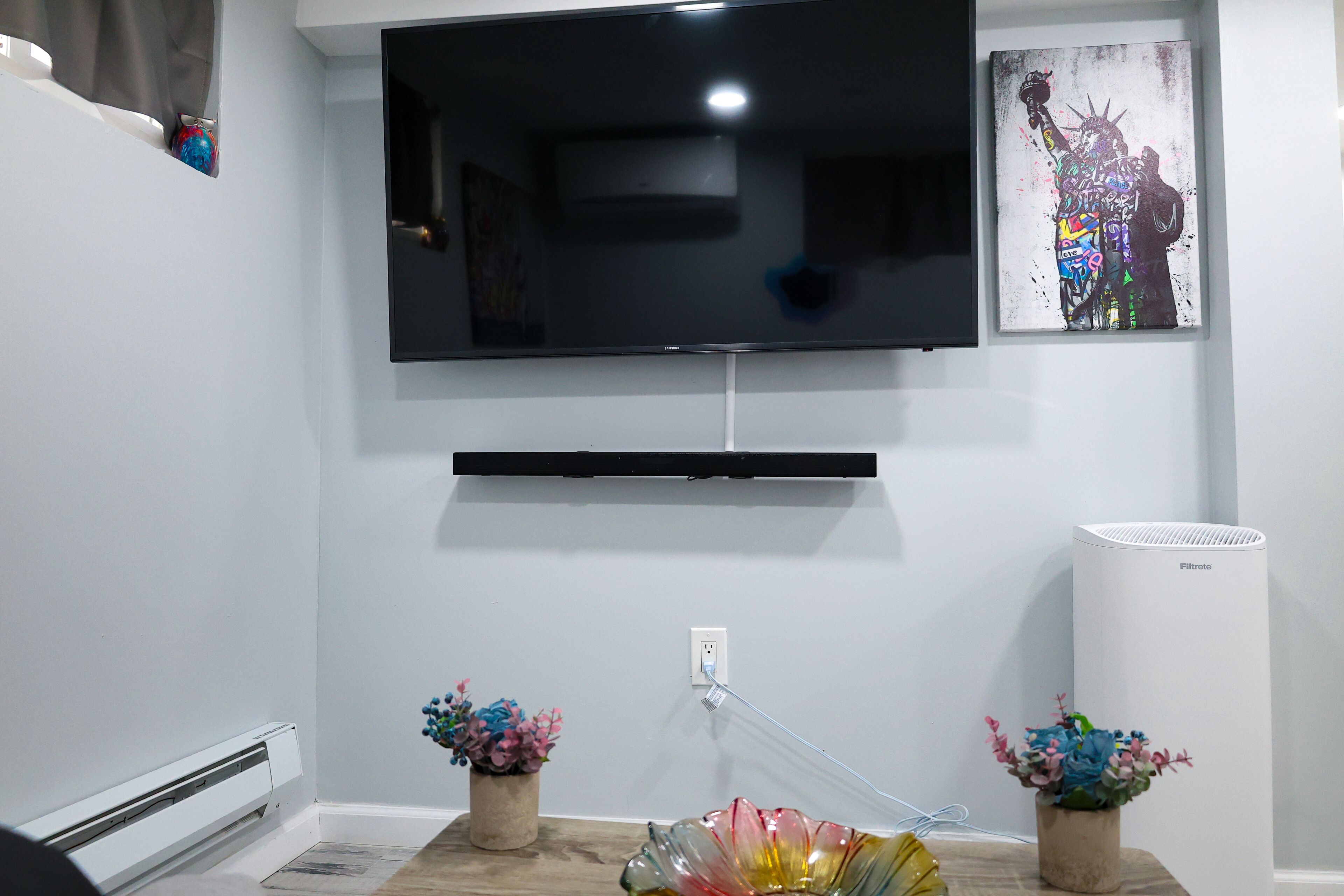 Een woonkamer met een wandgemonteerde flatscreen-tv, een witte airconditioner en twee decoratieve bloempotten op de vloer.