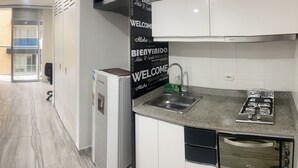 A compact hotel room with a bed, a small desk, a chair, a kitchenette with a sink and stove, and a sliding door leading to a balcony.