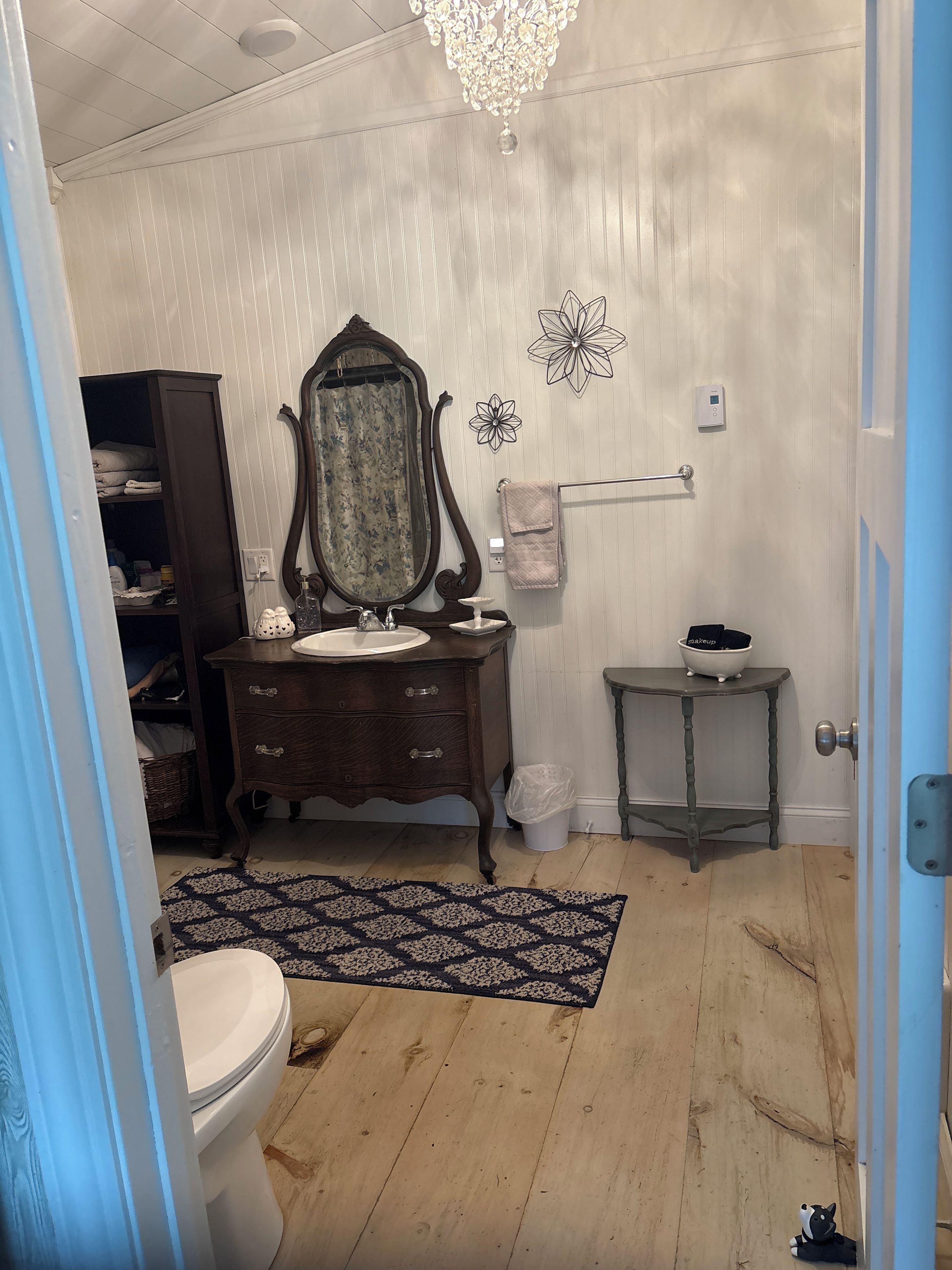 Une salle de bain avec un meuble-lavabo en bois, un grand miroir, des toilettes, une petite table avec un récipient pour les produits de maquillage et un élément décoratif mural.
