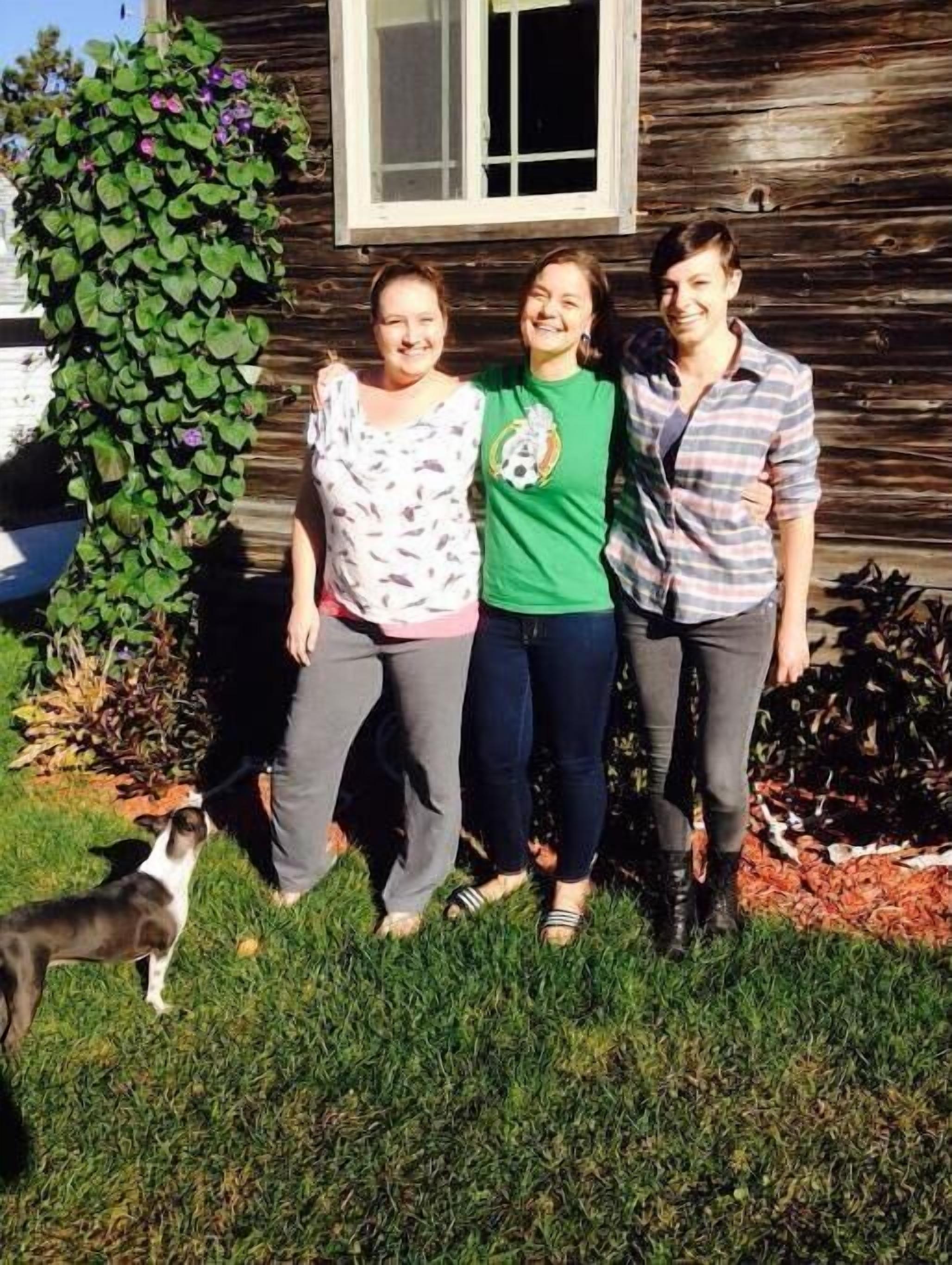 Three people standing in front of a wooden house with a window and a green plant.