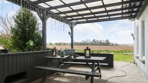 An outdoor patio with a wooden table and benches, surrounded by potted plants and a pergola.