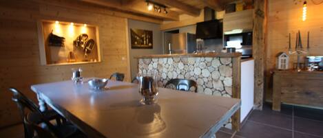 Une table de salle à manger en bois, des chaises, un mur en pierre et un coin cuisine équipé d’appareils électroménagers.