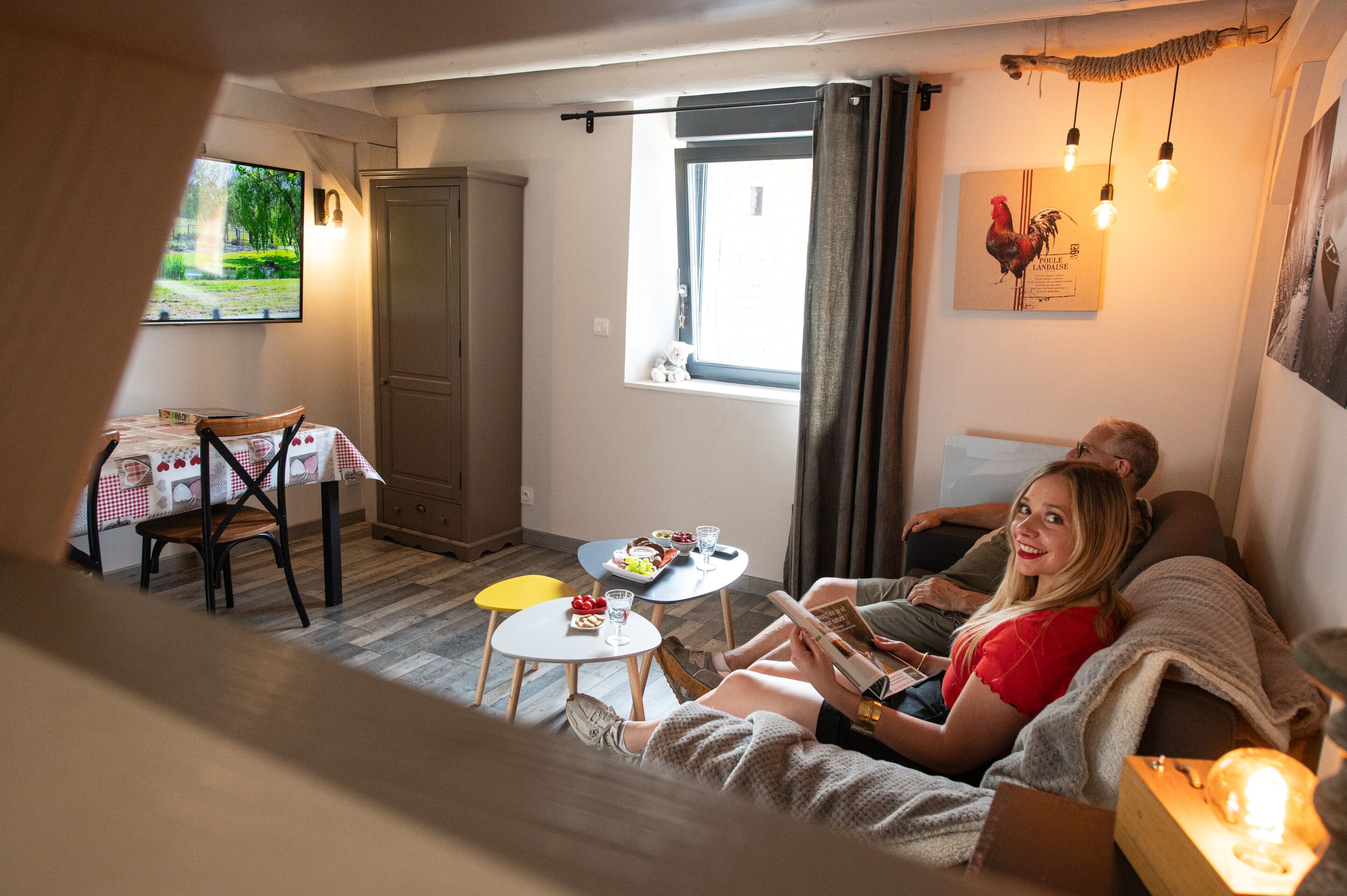 Un salon confortable avec un canapé, une table à manger et des chaises, une fenêtre avec des rideaux et un luminaire fixé au mur.