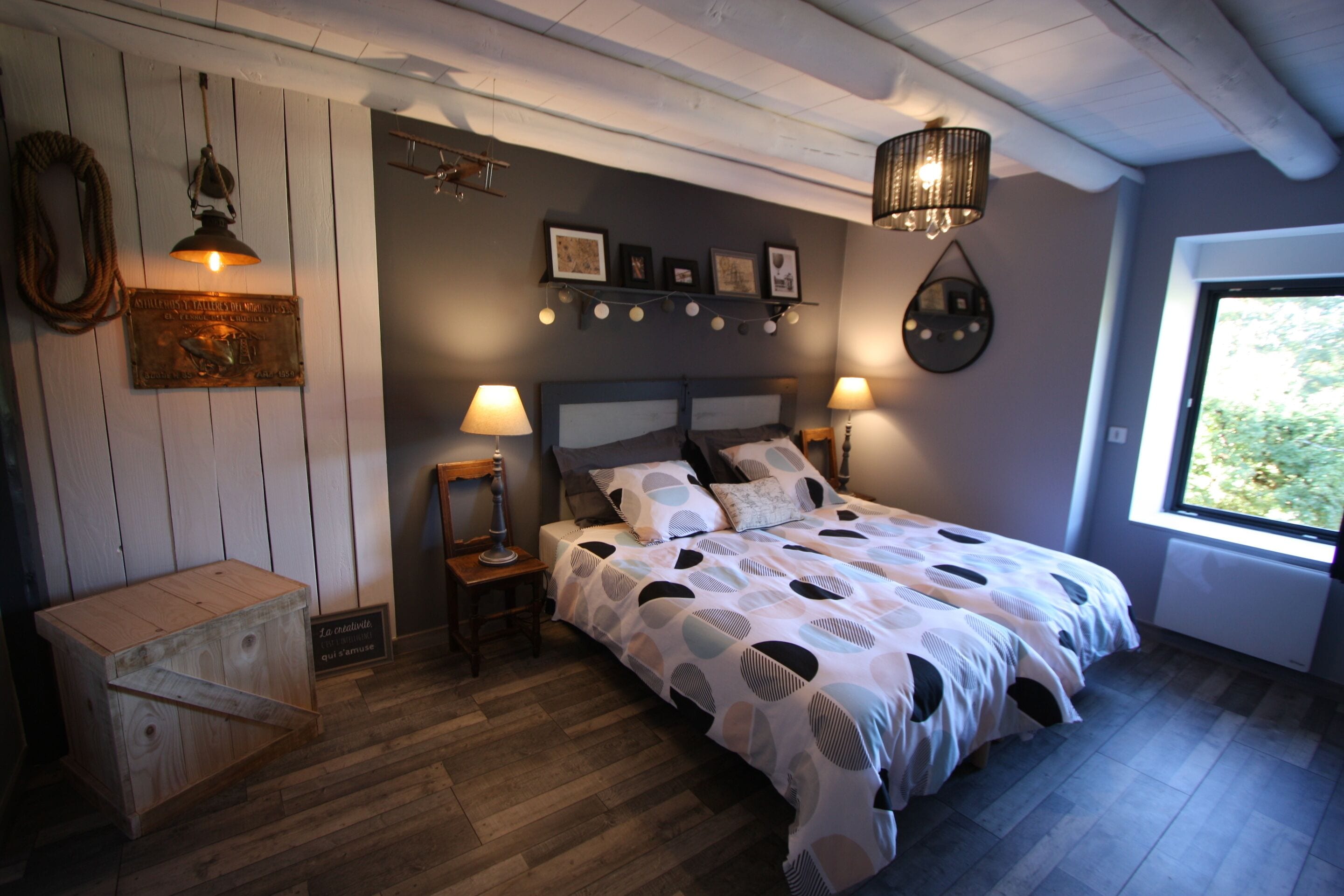 A bedroom with a bed, two bedside lamps, a wooden nightstand, a wall-mounted shelf with pictures, and a window with a view of greenery.