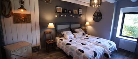 A bedroom with a bed, two bedside lamps, a wooden nightstand, a wall-mounted shelf with pictures, and a window with a view of greenery.