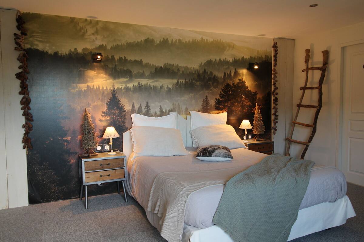 Ein Schlafzimmer mit einem großen Wandteppich mit Waldmotiv, einem Bett mit weißer Bettwäsche, zwei Nachttischen mit Lampen und einer an der Wand lehnten Holzleiter.