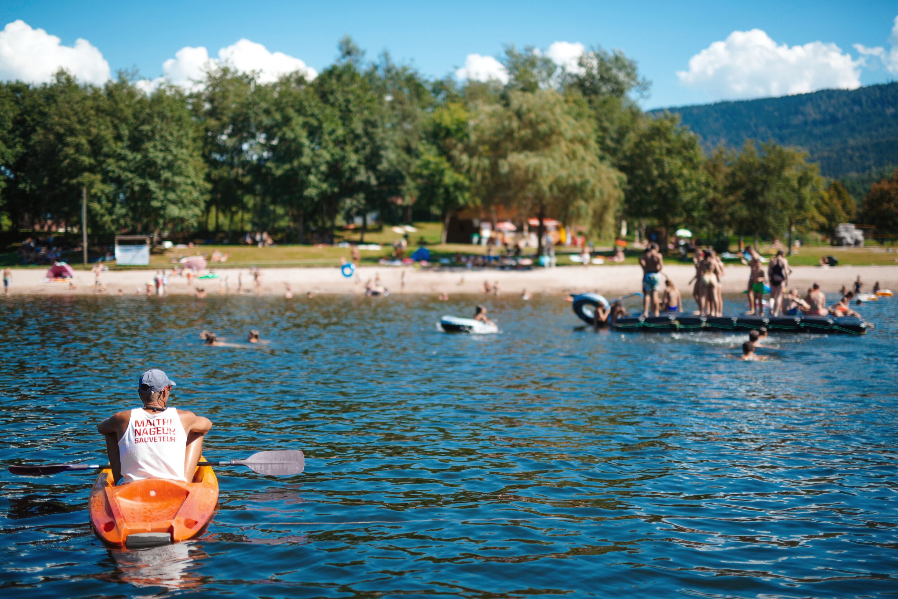 Un lago con gente en botes y en la orilla, rodeado de árboles y una montaña al fondo.