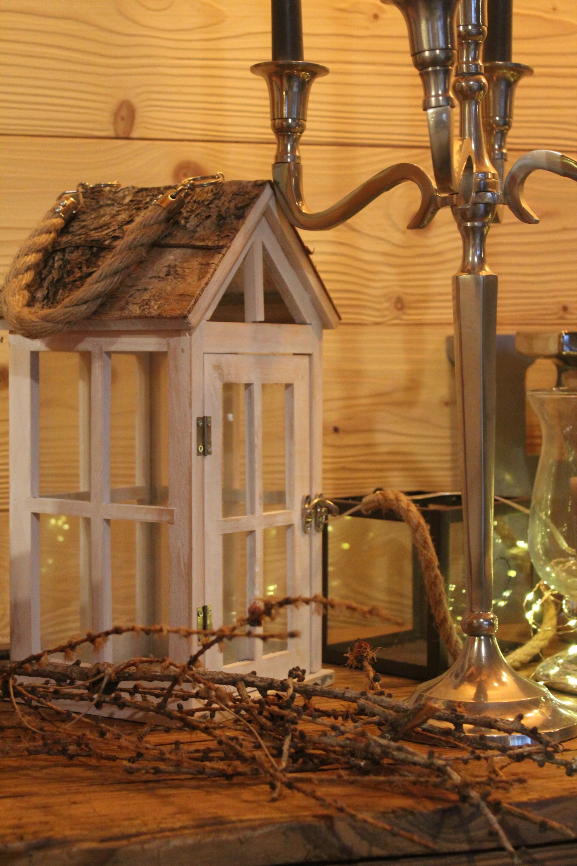 Un interior rústico de madera con un pequeño objeto en forma de casa blanca, un candelabro metálico alto y un florero de vidrio con una planta.