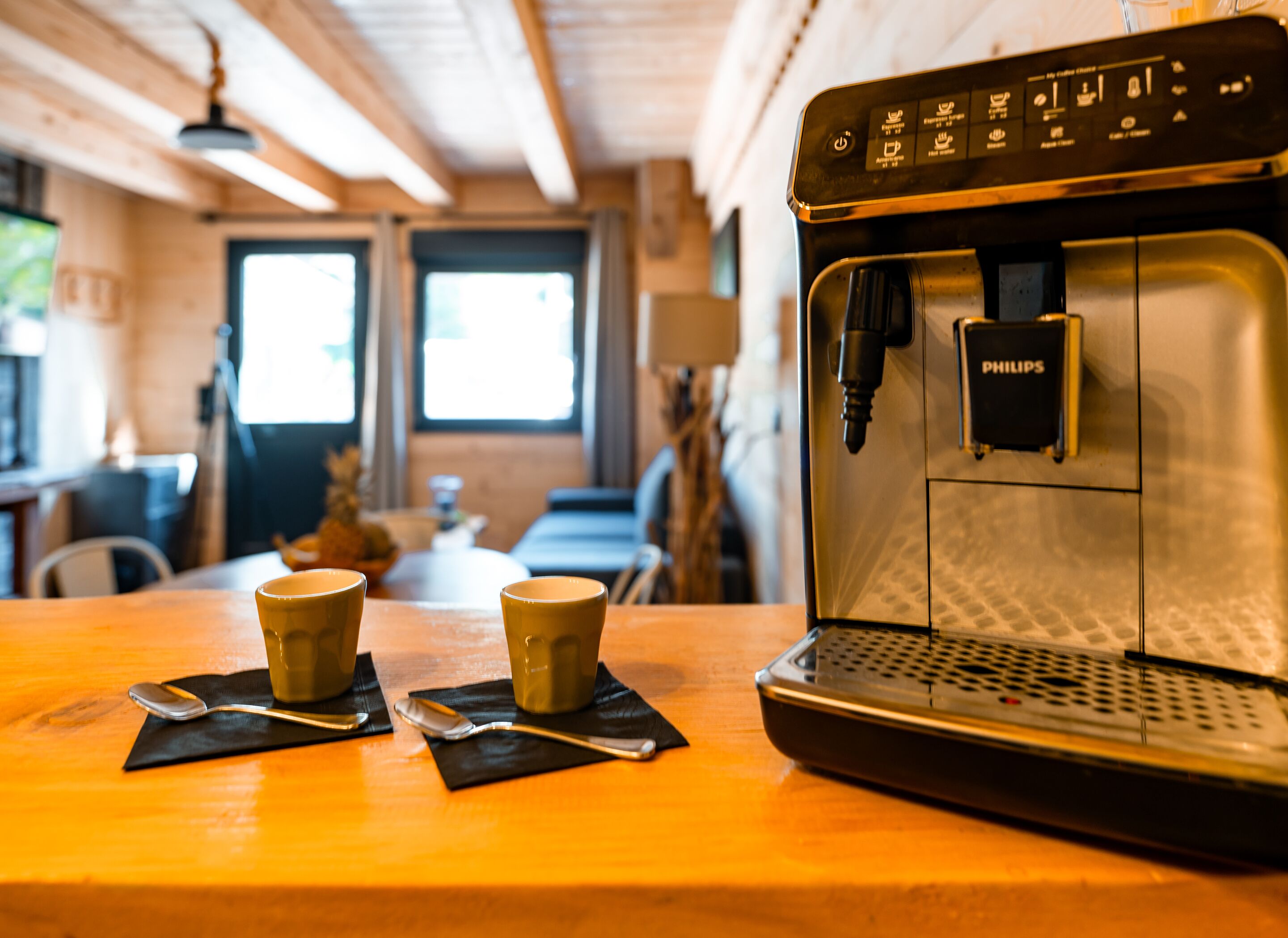 Une machine à café ornée du logo Philips, deux tasses et des cuillères, posés sur une table en bois dans une pièce dont le plafond est décoré de poutres en bois.