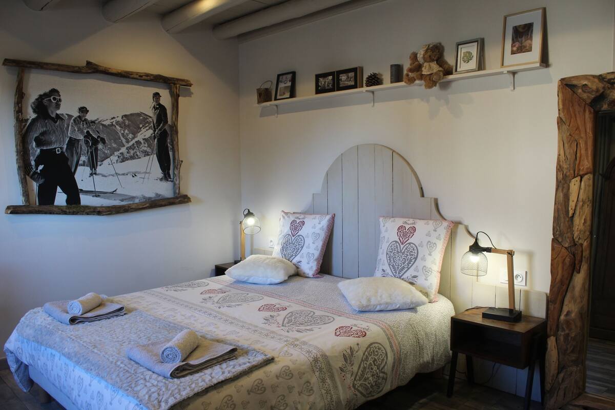 Ein ordentlich bezogenes Bett mit Bettwäsche mit Herzmuster, ein Wandbild von Skifahrern und ein Regal mit gerahmten Fotos und einem Teddybär.