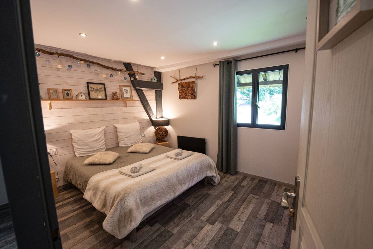 Ein Schlafzimmer mit einem Bett, einem Nachttisch, einer an der Wand befestigten Ablage mit Dekorationen, einem Fenster mit Vorhängen und einem Holzboden.