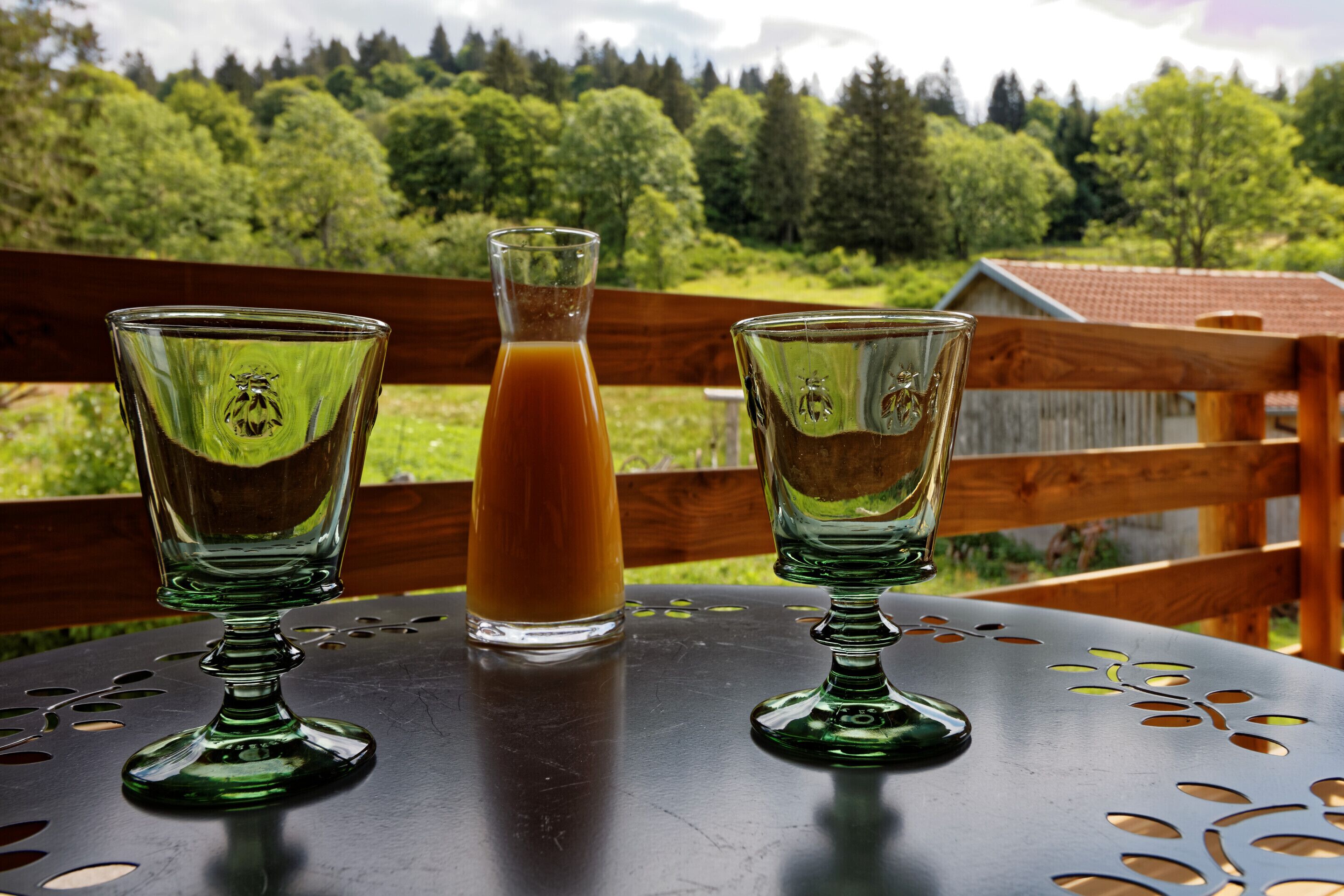 Dos vasos verdes con una botella de líquido ámbar sobre una mesa con vista a una valla de madera y árboles.