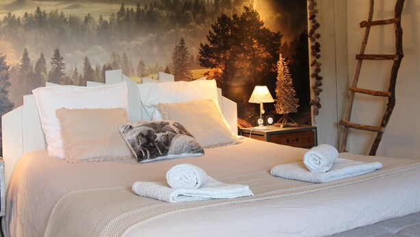 Une chambre à coucher avec un lit, des oreillers, une table de chevet avec une lampe, une échelle et une fresque représentant une forêt sur le mur.