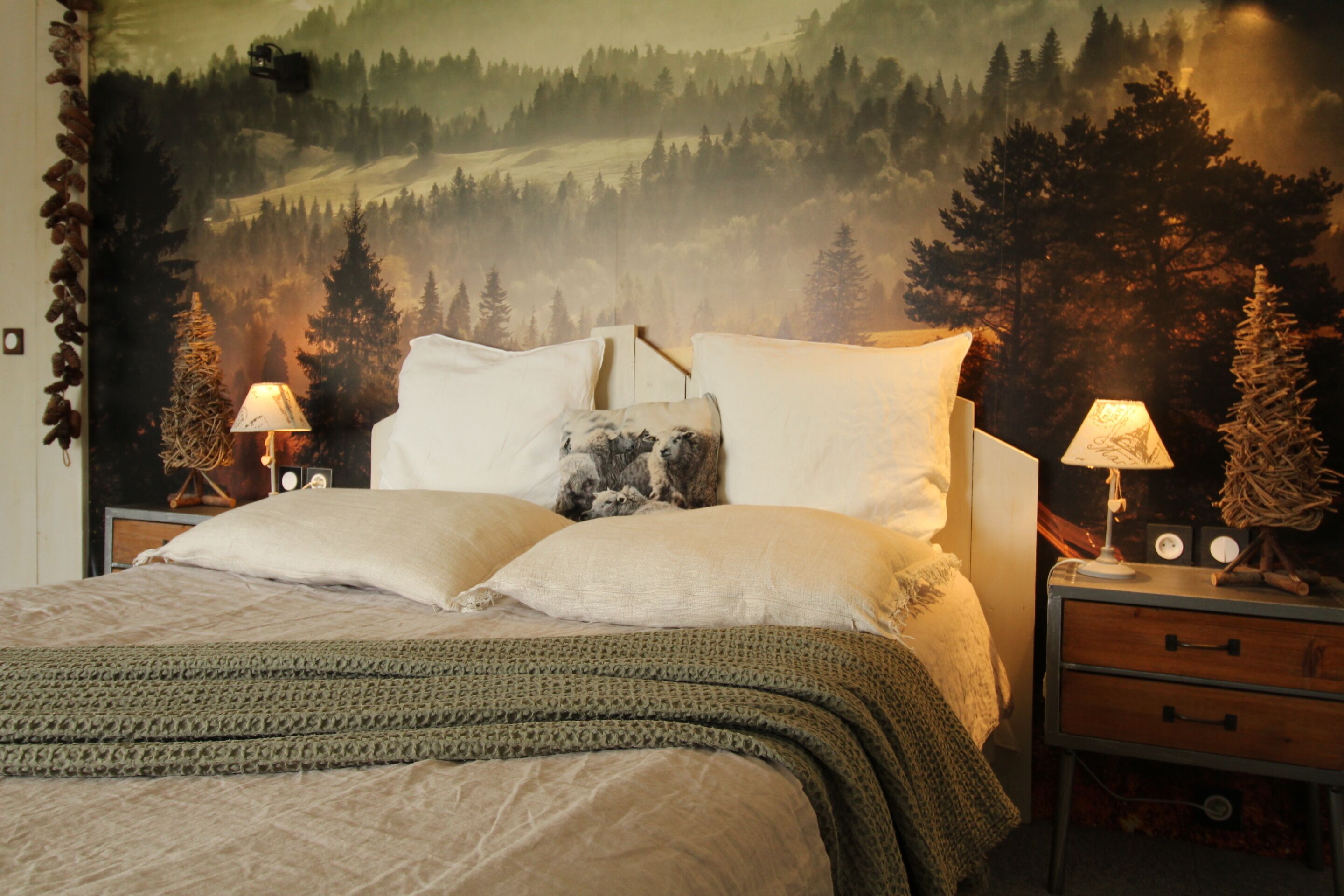 Un dormitorio con una cama, dos mesitas de noche, una lámpara y un tapiz que representa un paisaje forestal.