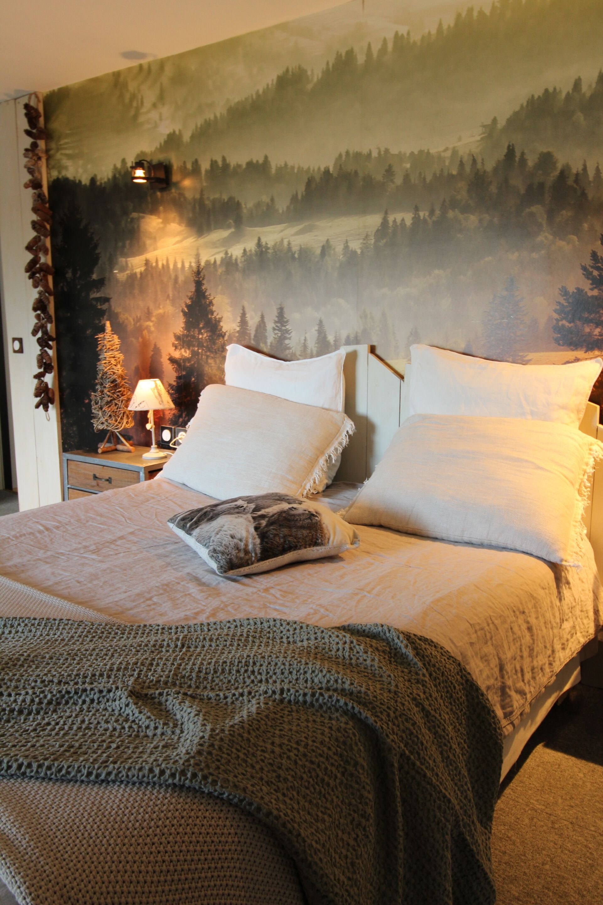 A bedroom with a bed, pillows, a blanket, a nightstand with a lamp, and a forest-themed wall mural.