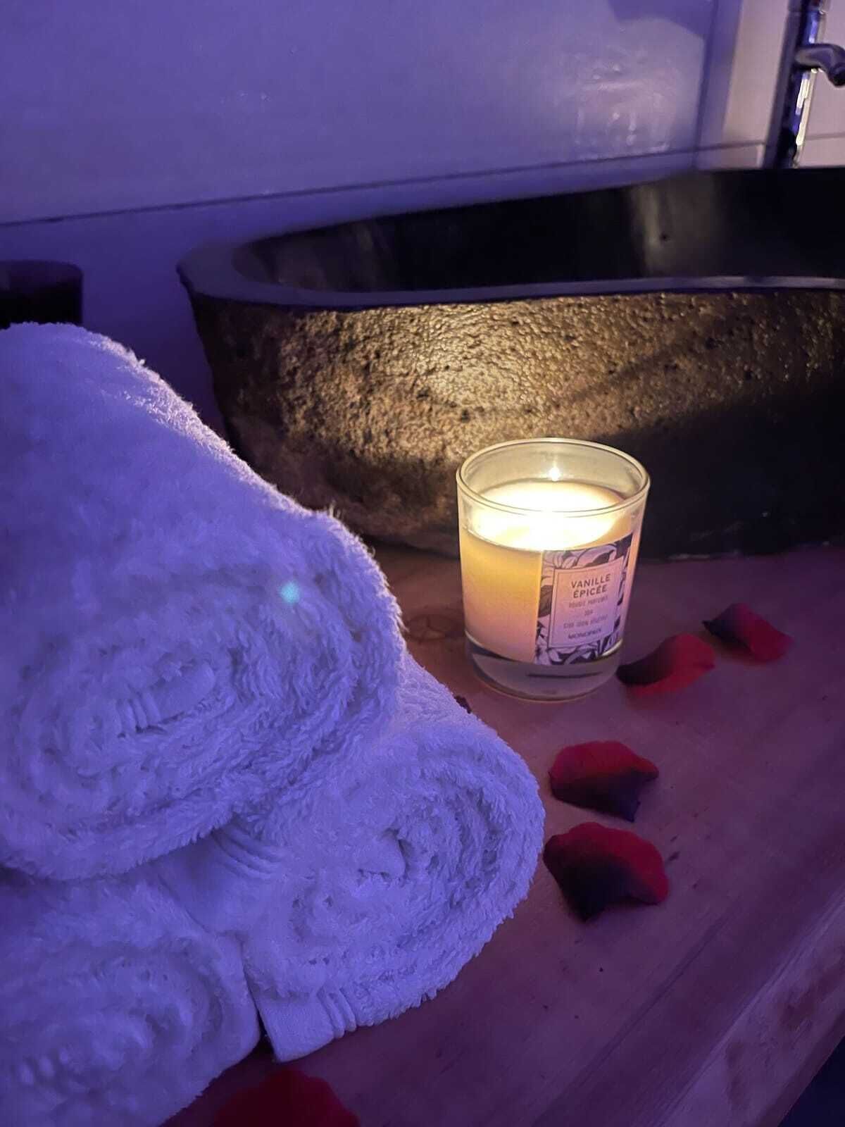 Une bougie, une serviette blanche enroulée et une baignoire remplie de pétales de roses, le tout posé sur une surface en bois.