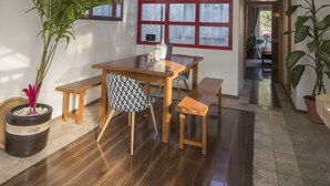 A dining area with a wooden table and benches, a large window with red frames, and potted plants.