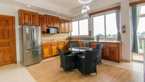 A modern kitchen with wooden cabinets, a stainless steel refrigerator, a dining table with chairs, and a large window providing a view of the outdoors.