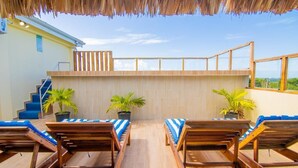 A rooftop terrace with wooden lounge chairs and potted plants, a wooden railing, and a view of the sea.