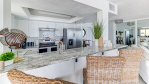 A modern kitchen with white cabinets, stainless steel appliances, and a marble countertop. There are wicker chairs and a potted plant.