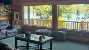 A cabin interior with a large window overlooking a lake, a plush sofa, and a wooden deck. - Christy's Cottage, Waterfront w/slide & Ice Shanty (Kalispell)