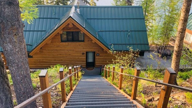 A wooden cabin with a metal roof, a staircase leading to the entrance, and a view of a lake and forest in the background. - Christy's Cottage, Waterfront w/slide & Ice Shanty (Kalispell)