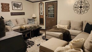 A living room with a sofa, ottoman, coffee table, decorative wall art, and a door with a decorative panel. - Animal Village/W/Mountain Views (Kalispell)