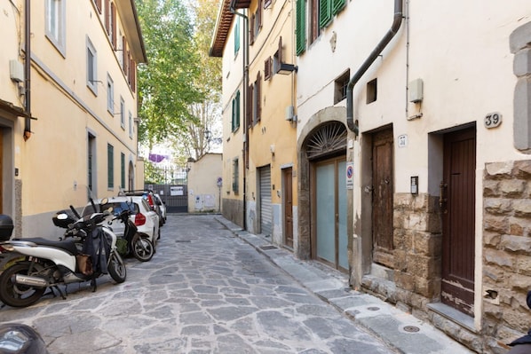 A narrow street with parked motorcycles and cars, stone pavement, and buildings with numbered doors and windows.