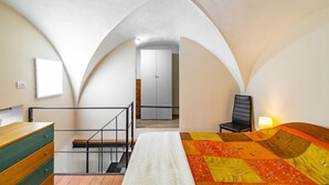 Una camera da letto con un letto, una sedia, una scrivania e un armadio. Si vede una scala che conduce a un'altra stanza.